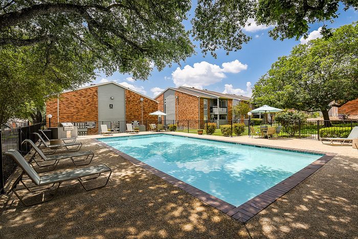Inviting apartment complex showcasing a sunny outdoor swimming pool with comfortable lounge chairs, surrounded by lush trees and modern brick buildings.
