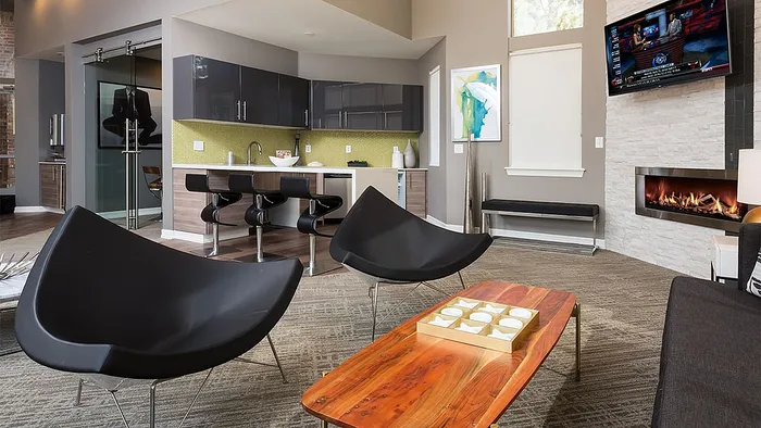 Modern living room with sleek black furniture, vibrant wood coffee table, and an elegant fireplace. Open kitchen with chic bar stools and stylish cabinetry enhance the contemporary vibe.