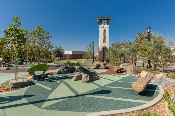 Charming outdoor park featuring a modern clock tower, lush greenery, and artistic rock formations under a clear blue sky.