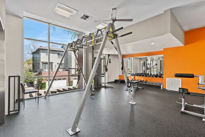 Modern fitness center with state-of-the-art equipment, vibrant orange accents, large windows with garden views, and ample natural light.