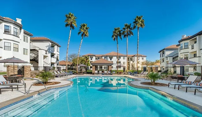 Luxurious apartment complex with a sparkling blue swimming pool, surrounded by elegant lounge chairs and towering palm trees under a clear blue sky.