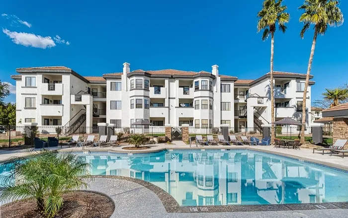 Luxurious apartment complex with modern architecture, surrounded by palm trees, featuring a sparkling pool with lounge chairs under a clear blue sky.