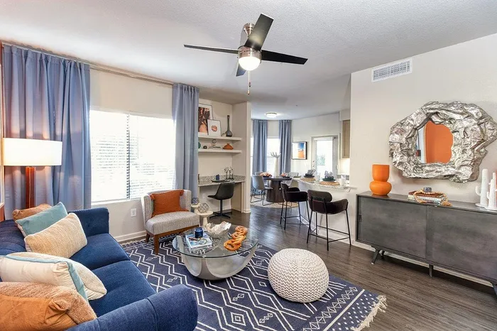 Bright and modern living area featuring a cozy blue sofa, stylish geometric rug, chic accent seating, and an elegant open kitchen with bar stools and contemporary decor.