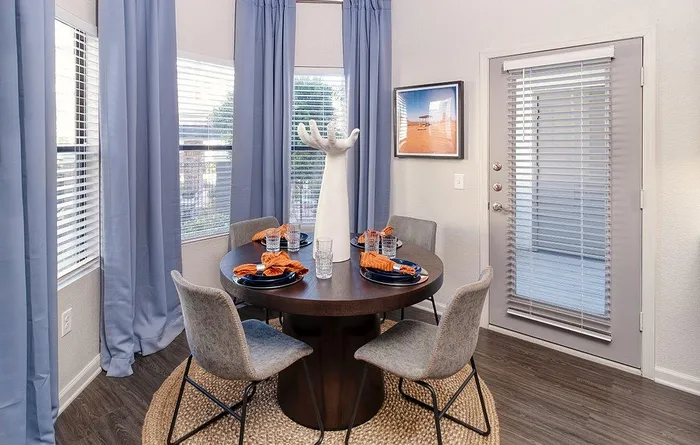 Chic dining area with a round table, stylish gray chairs, and bright blue-and-orange table settings, accented by elegant blue curtains and abundant natural light.