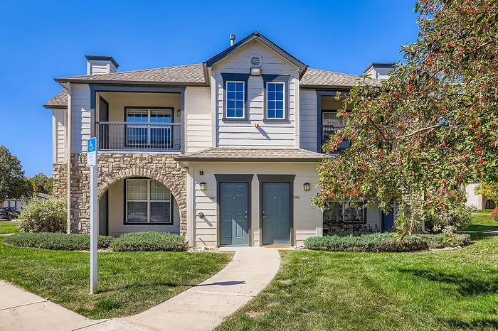 Charming two-story apartment building with stone accents, surrounded by lush greenery and a scenic blue sky backdrop.