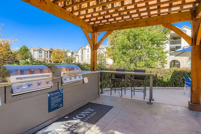 Inviting outdoor space with modern grills under a wooden pergola, surrounded by lush greenery and elegant apartment buildings in the background.