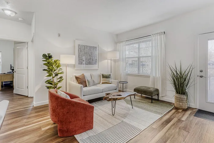 Bright and inviting living room with modern decor, featuring a cozy white sofa, vibrant rust-colored chair, and natural wood coffee table on stylish rugs.