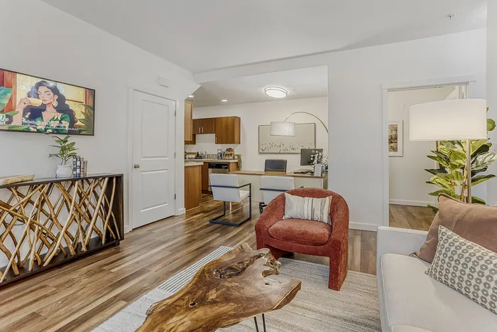 Inviting living space with stylish decor featuring a plush armchair, unique wooden coffee table, and open view into a modern kitchen and workspace.