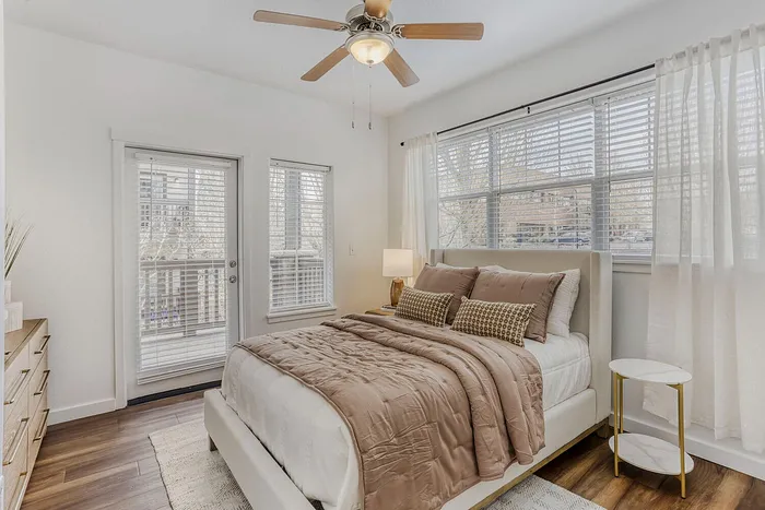 Inviting bedroom with elegant decor, featuring large windows with blinds, a plush bed with cozy bedding, and stylish wooden flooring.