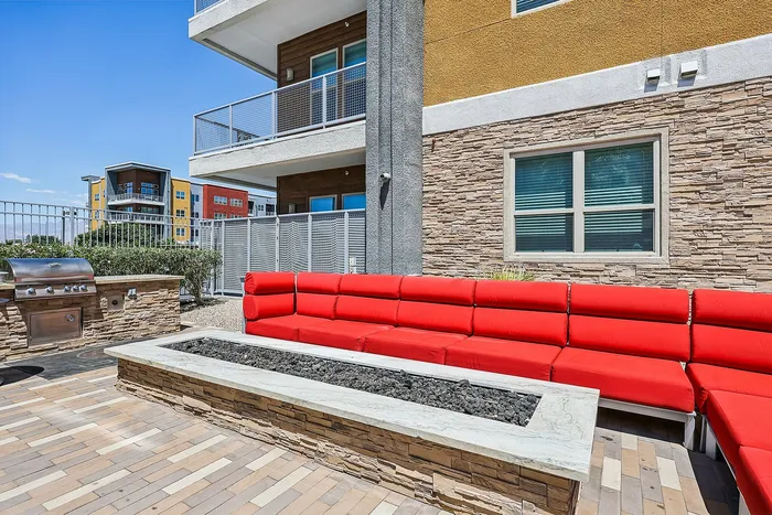 Modern outdoor patio with vibrant red seating, built-in stone barbecue grill, and sleek fire pit, set against a contemporary apartment building backdrop.