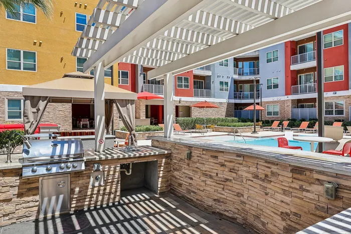 Modern apartment complex with vibrant colors featuring a barbecue area, shaded seating, and a sparkling pool surrounded by red umbrellas and lounge chairs.