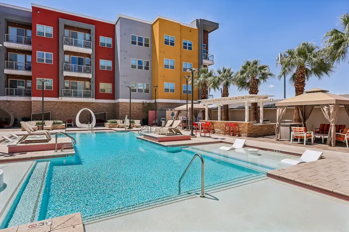 Vibrant apartment complex featuring a sparkling outdoor pool, stylish lounge chairs, and cozy cabanas under a bright blue sky.