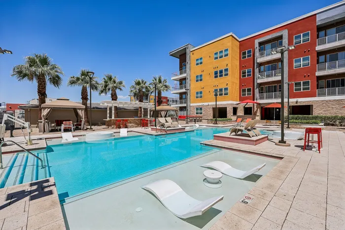 Vibrant apartment complex pool area featuring a luxurious swimming pool, stylish sun loungers, and surrounding palm trees against a bright blue sky.