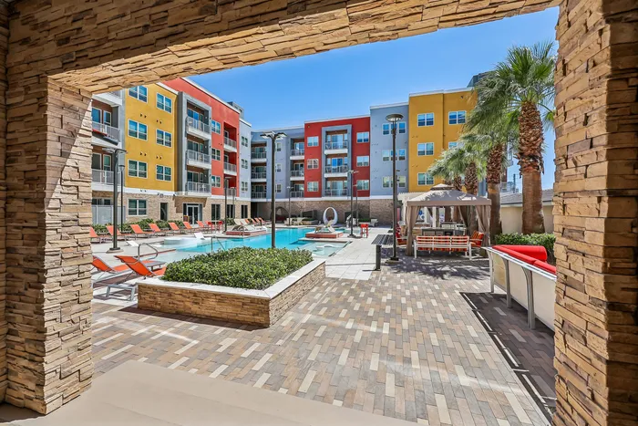 Vibrant apartment complex with a modern swimming pool area, colorful buildings, palm trees, and lounging spaces under a clear blue sky.