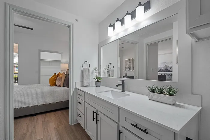 Model bathroom with white counters, white cabinets, large mirror and bedroom in the background