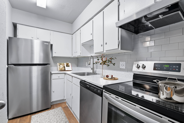 Renovated kitchen stainless-steel refrigerator, oven, and dishwasher, sleek white cabinets, gray subway tile backsplash, and wood flooring with gray rug