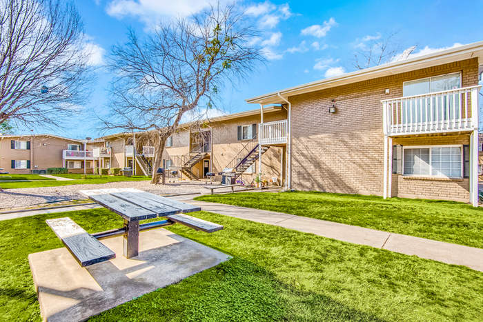 Exterior two story brick apartment with white patio fencing, grass, and treesExterior two story brick apartment with white patio fencing, grass, trees, and bench