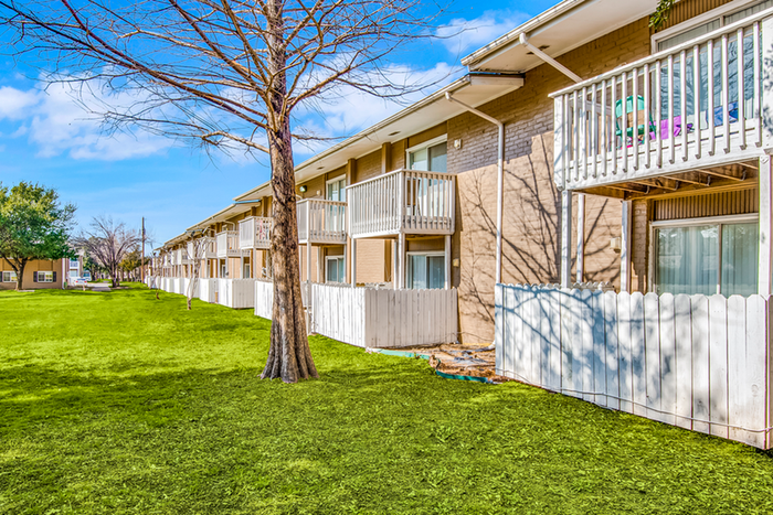 Exterior two story brick apartment with white patio fencing, grass, and trees