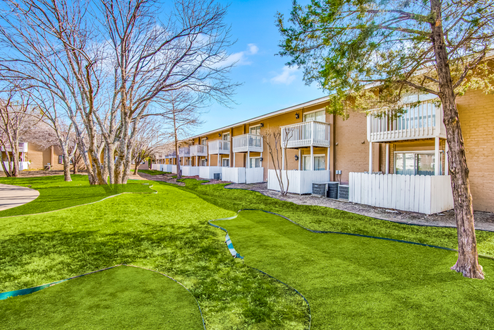 Exterior two story brick apartment with white patio fencing, grass, and trees