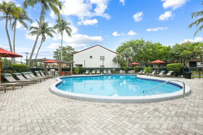 Pool with lounge chairs on sundeck, red umbrellas, palm trees and native landscaping with exterior building