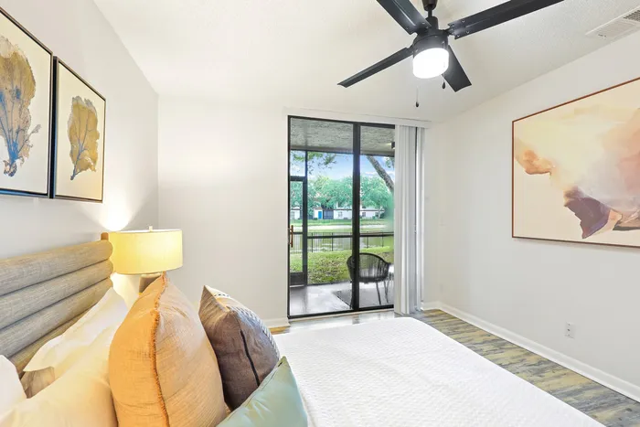 Bedroom with bed, decorative pillows, ceiling fan light fixture, floor to ceiling glass patio door, wall art