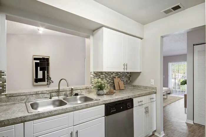 Kitchen with tan granite counters, white cabinetry, stainless steel dishwasher, mosiac backsplash, wall opening to living room
