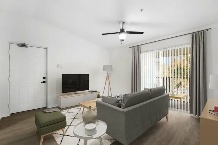 Living room with gray couch, green ottoman, hardwood style flooring with rug, glass patio doors with gray curtains, and a flat screen TV on entertainment center