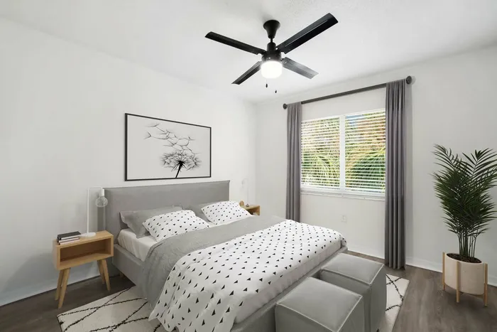 Bedroom with queen size bed with gray framing and bedding, wooden end table, hardwood style flooring with rug, large window with gray curtains