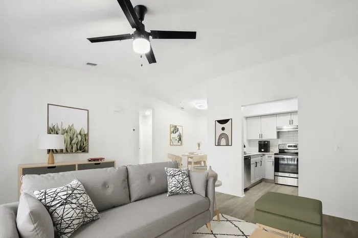 Living room gray couch, ceiling fan light fixture, hardwood style flooring, wall art, opening to kitchen stainless steel appliances
