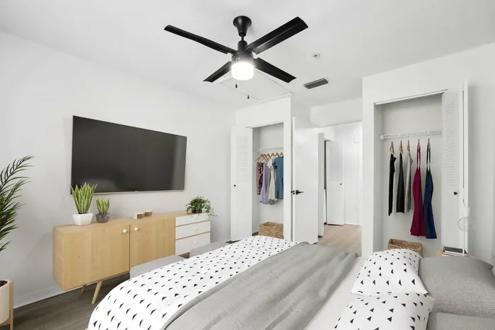 Bed with gray and white bedding, wood dresser, ceiling fan light fixture, closet to right of door and closet left of door, hardwood style flooring