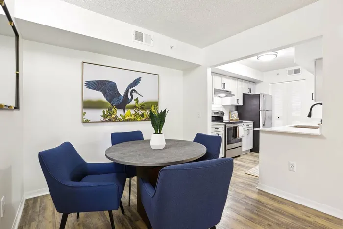 Dining area, round table, blue felt chairs, hardwood style flooring, opening to kitchen, stainless steel appliances, shaker counters, wall art