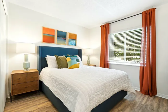 Bedroom, hard wood style floor, wooden end table, blue fabric bed framining, queen size bed with white bedding, large wind with orange curtains, wall art, table lamp