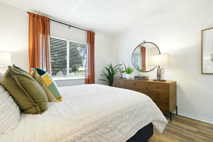 Hard wood style flooring, twin bed with white bedding, wooden dresser, circular wall mirror, table lamp, large window, orange curtains