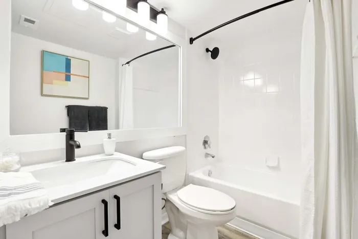 White quartz counters, shaker cabinets, vanity lighting and large mirror, toilet, bathtub, curved shower rod, wall art