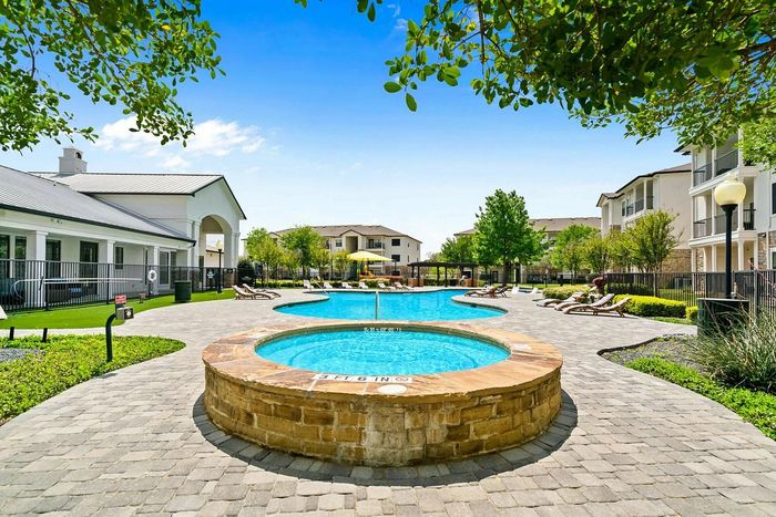 Luxurious apartment community featuring a sparkling outdoor pool with hot tub, surrounded by lush greenery and elegant lounging areas.