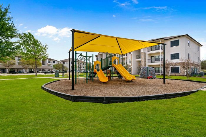 Vibrant community playground with yellow shade cover and slides, set in a lush garden area with modern apartment buildings in the background.