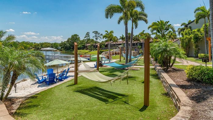 Lakeside oasis with inviting hammocks, vibrant blue Adirondack chairs, and lush tropical landscaping under a clear blue sky.