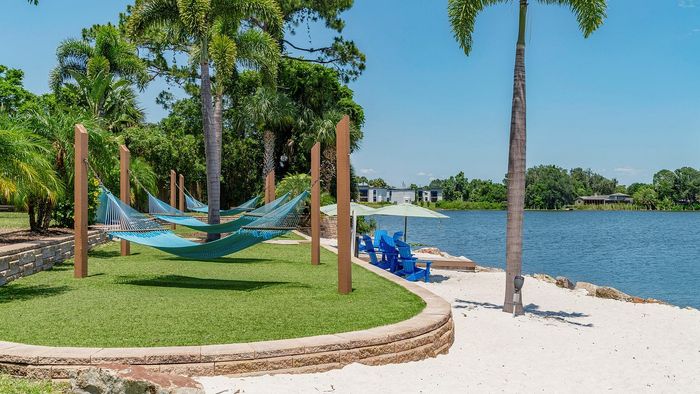 Serene lakeside setting with cozy blue hammocks and Adirondack chairs on sandy shores, surrounded by lush palm trees.