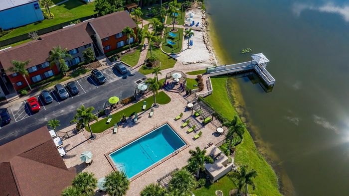 Breathtaking aerial view of a modern riverside apartment complex with a sparkling pool, lush green landscapes, and a scenic boardwalk.