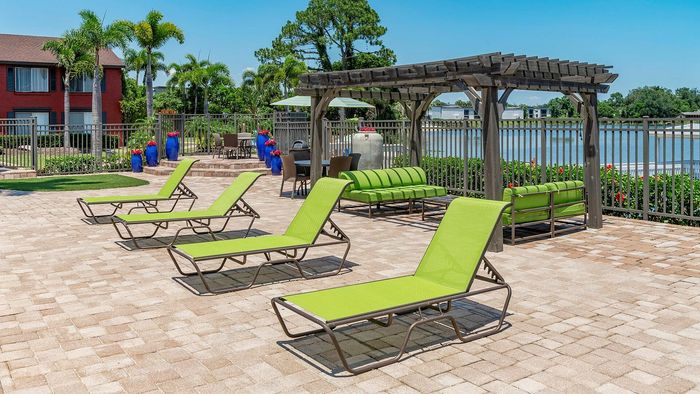 Luxurious poolside retreat with vibrant green lounge chairs, cozy outdoor seating under a stylish pergola, surrounded by lush palm trees and tranquil water views.