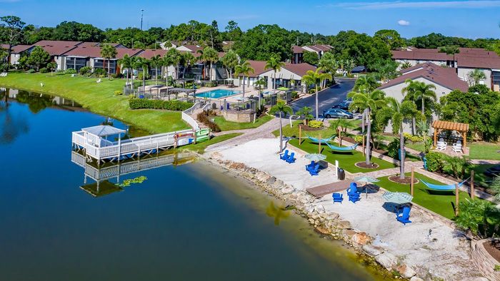 Scenic apartment complex with a tranquil lakeside view, featuring landscaped greenery, inviting pool area, and vibrant beach chairs under palm trees.