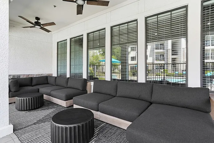 Elegant outdoor lounge area with modern grey sectional sofas, stylish ceiling fans, and large windows offering a view of the pool.