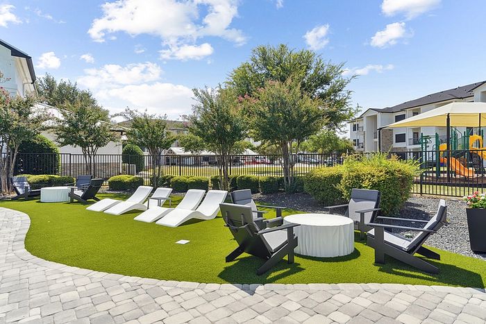 Inviting outdoor space with modern lounge chairs, vibrant greenery, and a cozy seating area set against a backdrop of stylish apartment buildings and a playful children's playground.