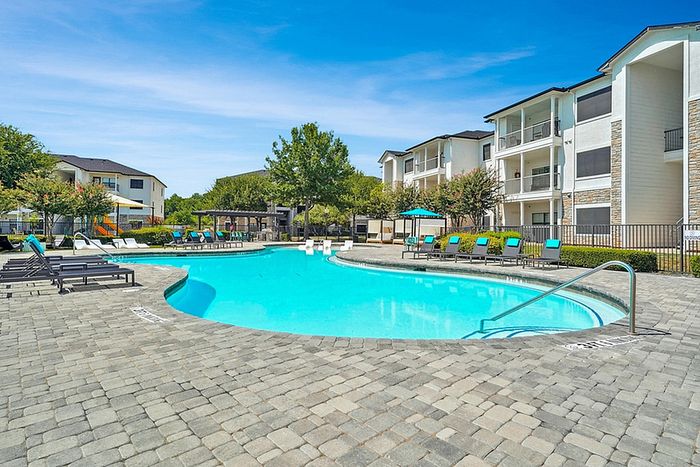 Luxurious apartment community with a swimming pool, lined with sun loungers, surrounded by lush greenery and modern building exteriors under a clear blue sky.