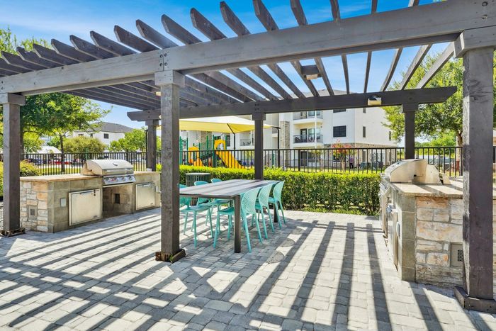 Inviting outdoor community space featuring a stylish pergola with modern barbecue grills and vibrant teal seating, offering a perfect socializing area next to a playground and lush greenery.