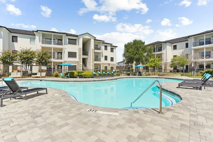 Chic apartment community featuring a pool surrounded by modern lounge chairs and lush greenery, under a bright blue sky.