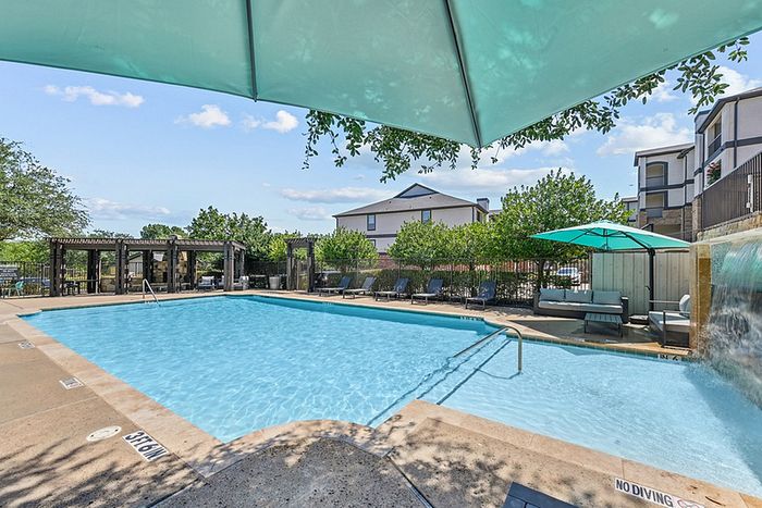 Luxurious apartment community pool with refreshing water features, shaded lounge chairs, and vibrant umbrellas creating a relaxing oasis.