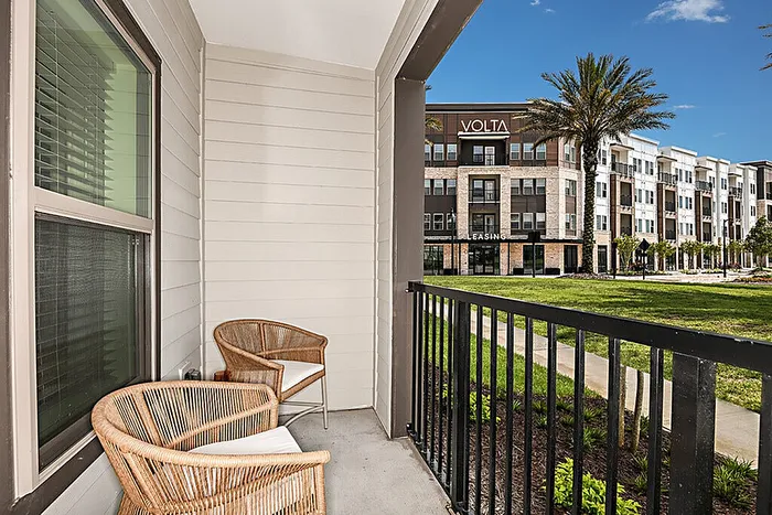 Charming apartment patio with wicker chairs overlooking lush green space and elegant modern building.