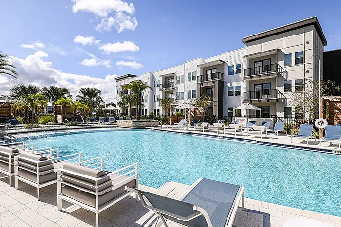 Luxurious apartment community showcasing a outdoor swimming pool, chic poolside seating, and elegant four-story buildings under a brilliant blue sky with palm trees creating a serene tropical atmosphere.