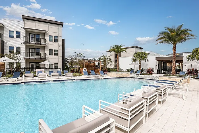 Luxurious apartment community featuring a swimming pool area with modern lounge chairs, surrounded by lush palm trees and stylish architecture under a bright blue sky.